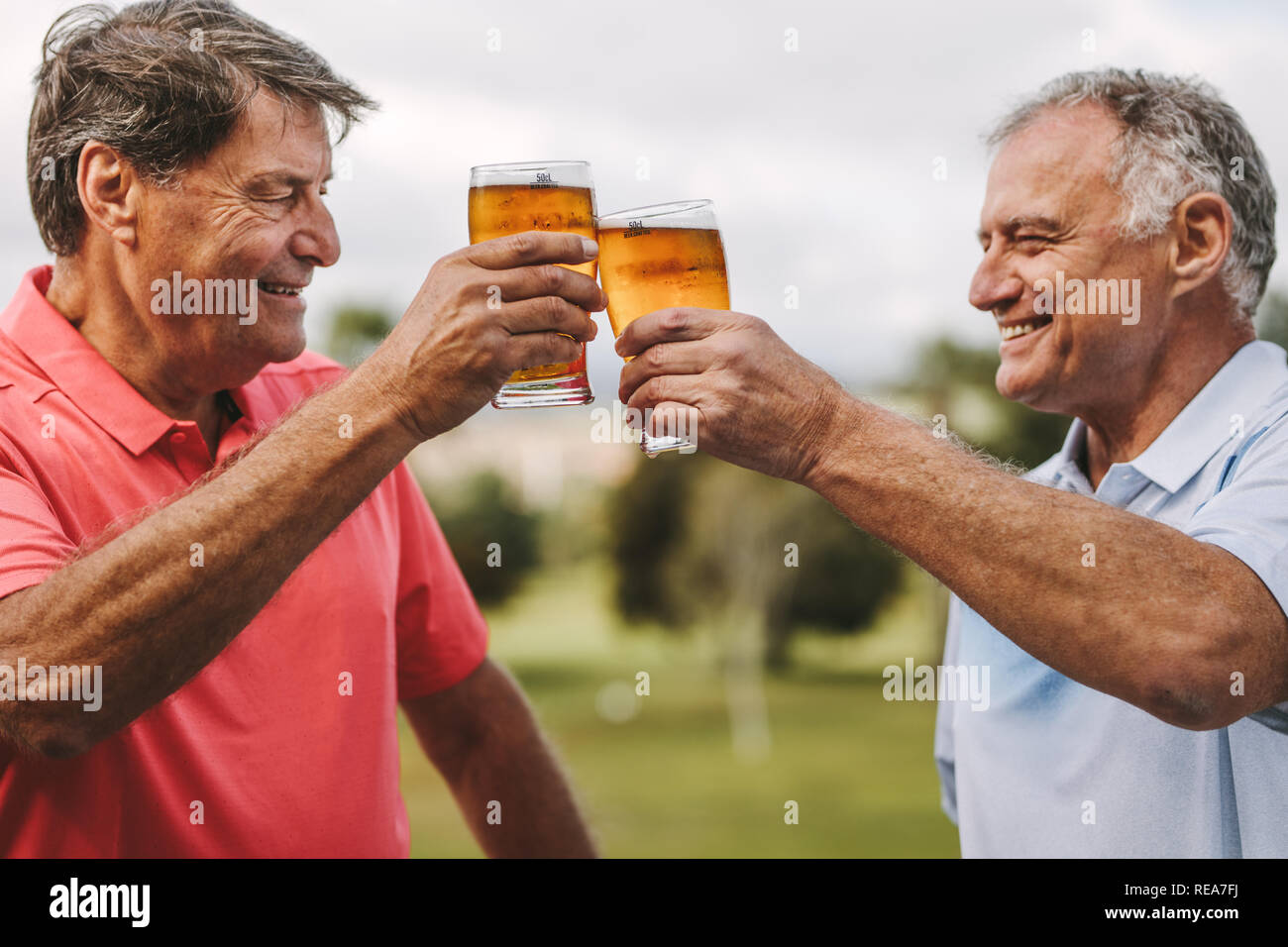 Two senior men toasting beer glasses outdoors. Smiling mature male friends cheering beers while standing outside. Stock Photo