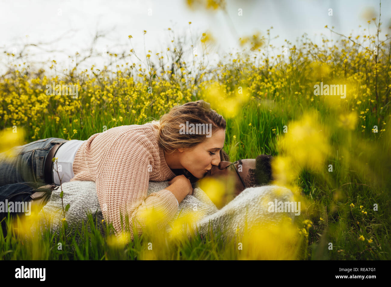 Romantic interracial couple lying in green grass and kissing. Loving couple kissing on flowering meadow outdoors. Stock Photo
