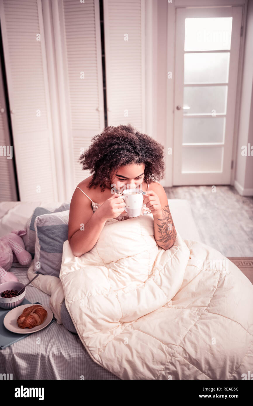 Joyful young lady sitting in her bed and drinking freshly-made coffee Stock Photo
