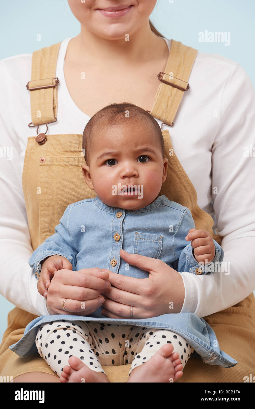 Cropped portrait of adorable little baby girl in jeans dress looking at camera in her mother s arms against pale blue background Stock Photo Alamy