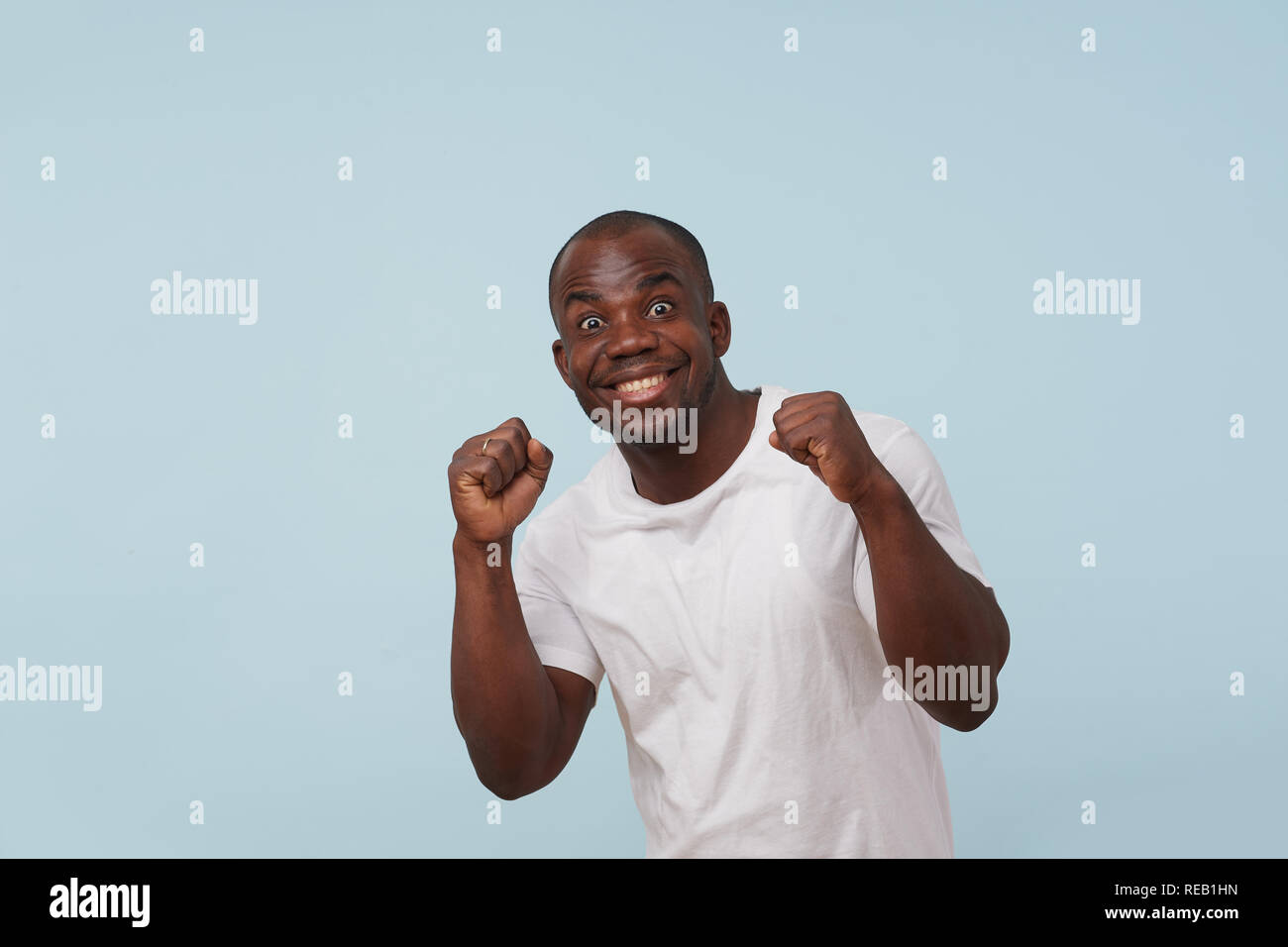 Handsome bold black man in white T-shirt is looking at camera and smiling,  grimacing against pale blue background. Fists closed, crazy eyes wide open  Stock Photo - Alamy