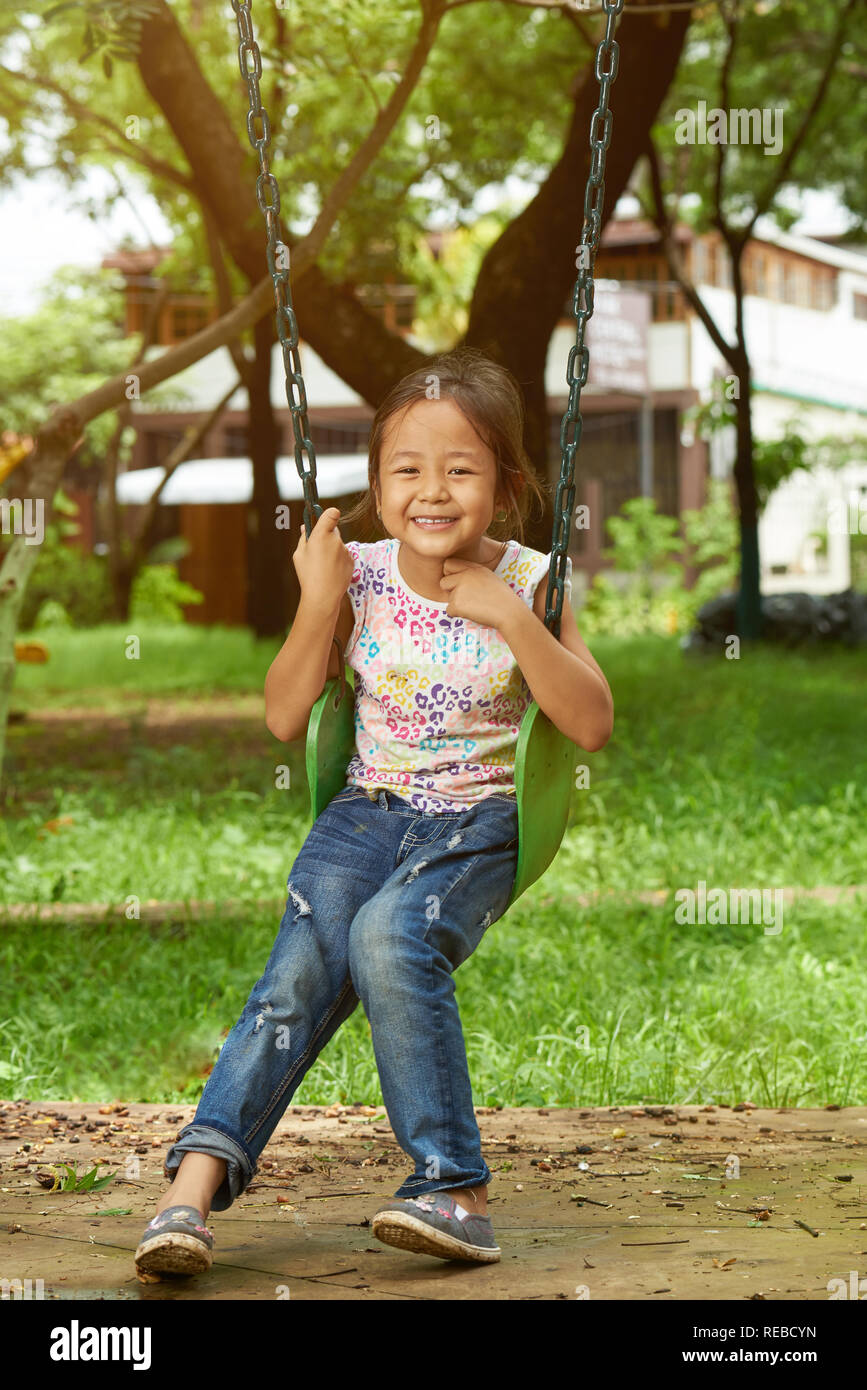 Smiling asian girl sitting on swings in sunny bright day background Stock Photo
