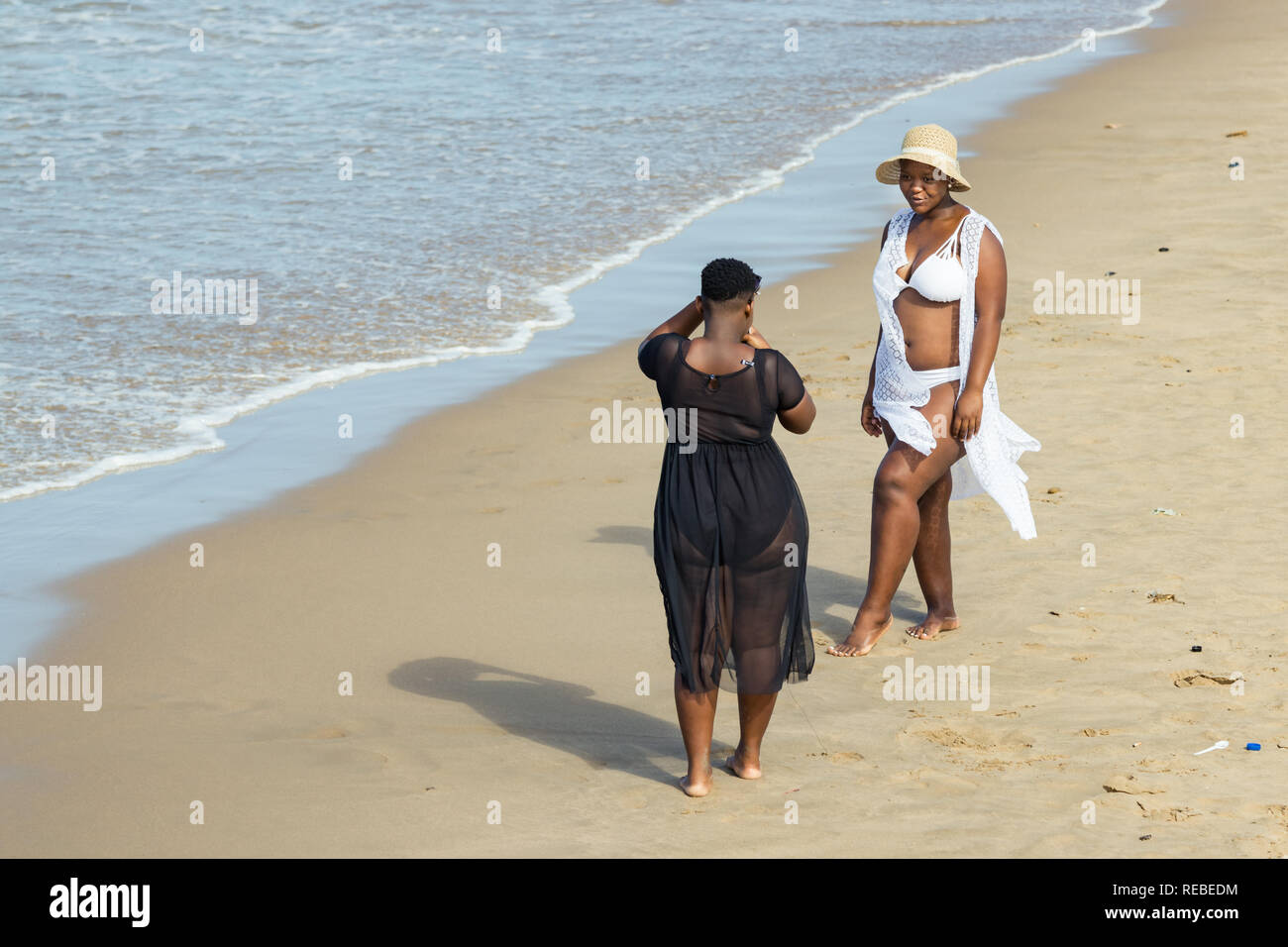An oversize tall black woman wearing a white bikini and hat posing for her friend who takes her photos in the beach in Durban, South Africa. Stock Photo