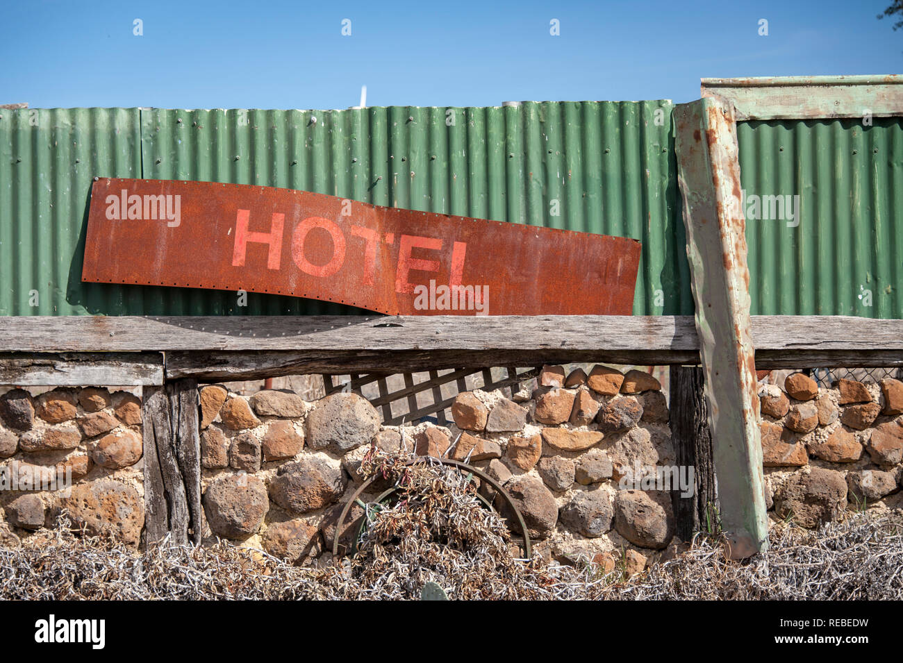 Old and rusted disused hotel sign leaning against a green corrugated fence on top of a stone wall Stock Photo