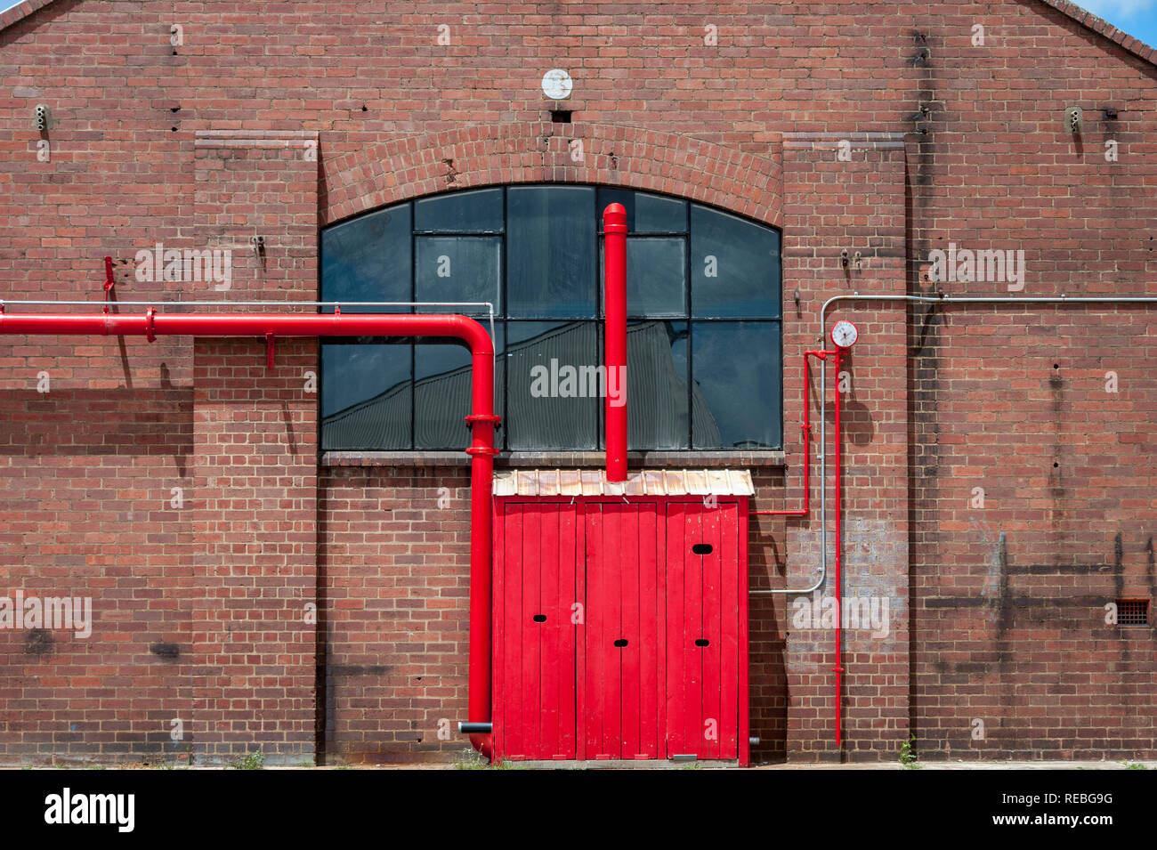 A red control box and red pipes against a brick building with arched windows Stock Photo