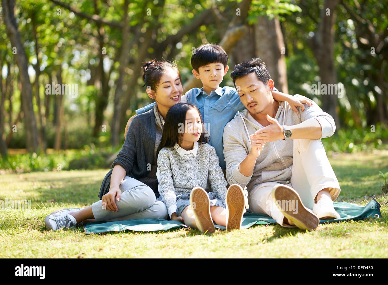 asian family with two children having fun sitting on grass talking chatting outdoors in park Stock Photo