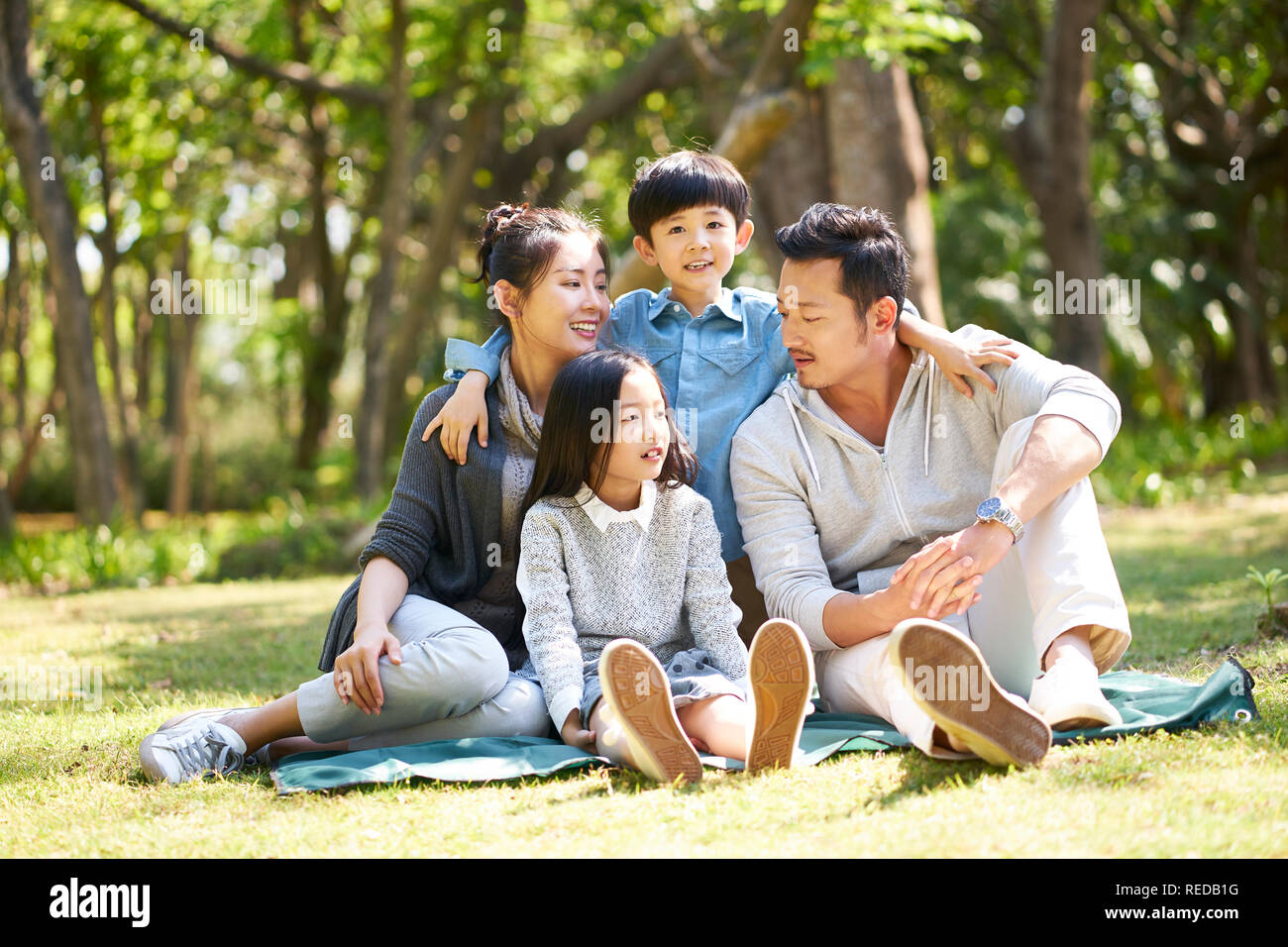 asian family with two children having fun sitting on grass talking chatting outdoors in park Stock Photo