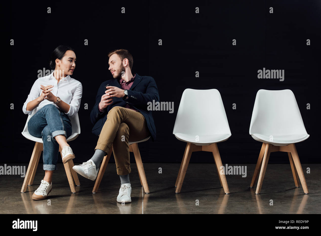 multiethnic couple sitting on chairs and talking isolated on black Stock Photo