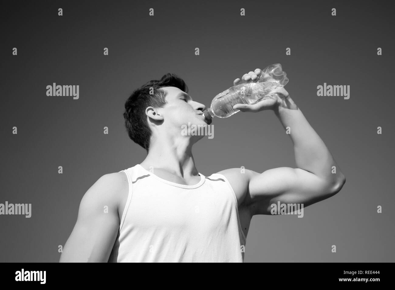 Bodybuilder with muscular hands muscles biceps, triceps. Athlete in green tshirt on blue sky. Sport and fitness concept. Healthy lifestyle and health. Man drinking water from bottle on sunny day. Stock Photo