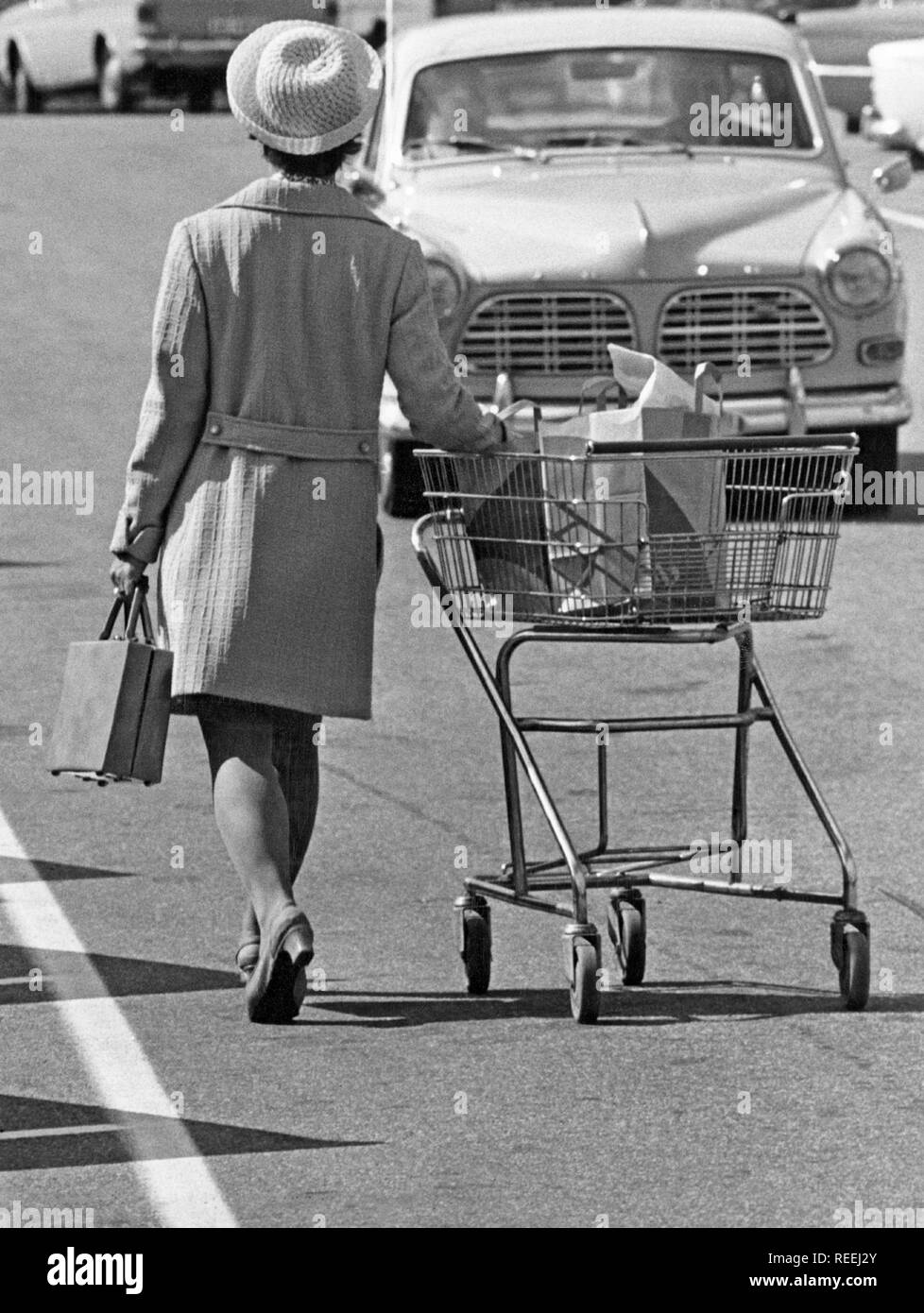 Shopping in the 1970s. A lady is leaving the supermarket and heading for her car pushing the shopping cart on the parking lot. Sweden 1970 Stock Photo
