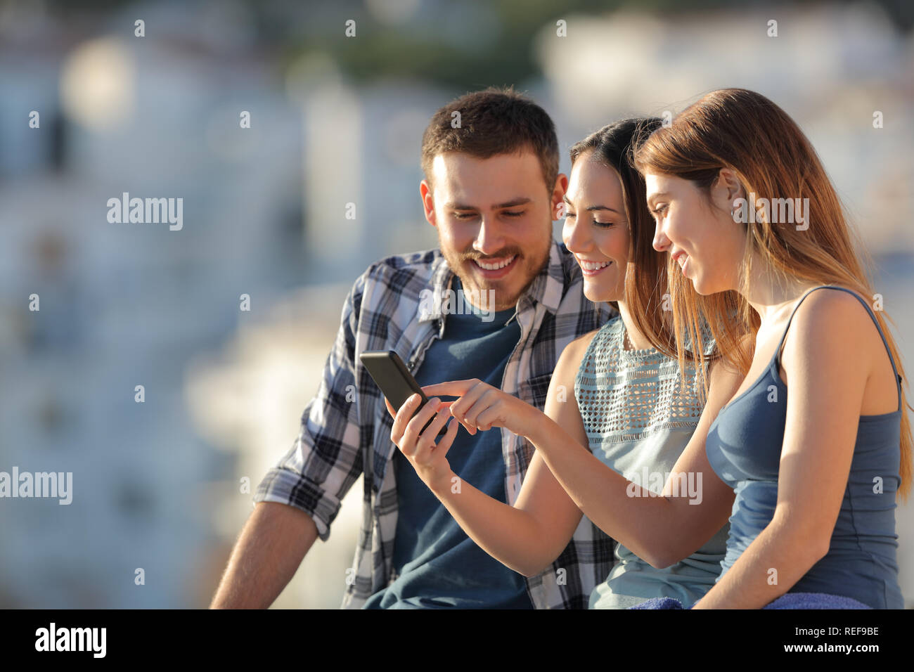 Three happy friends using a smart phone in a town at sunset Stock Photo