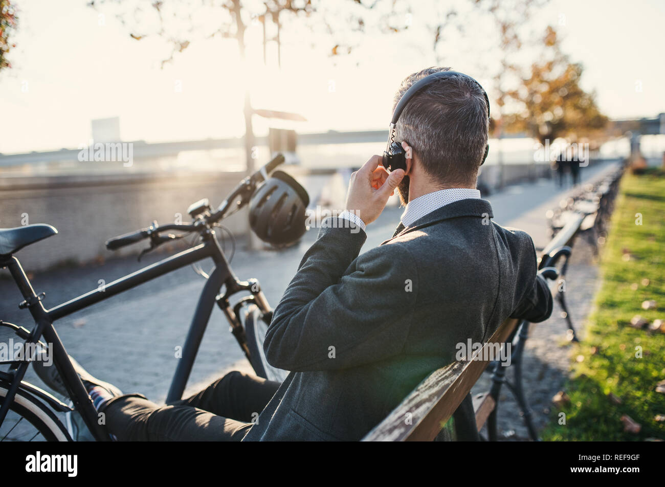 Businessman commuter with bicycle sitting on bench in city, listening to music. Stock Photo