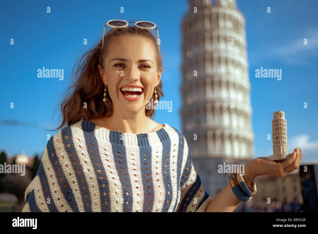 smiling elegant woman in striped blouse in the front of leaning tower in Pisa, Italy showing leaning tower of Pisa souvenir. сaucasian woman with brow Stock Photo