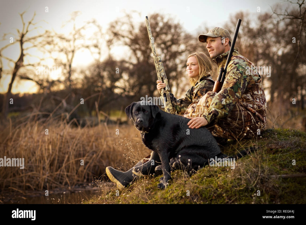 Young adult couple duck shooting with their dog by a lake at sunset. Stock Photo