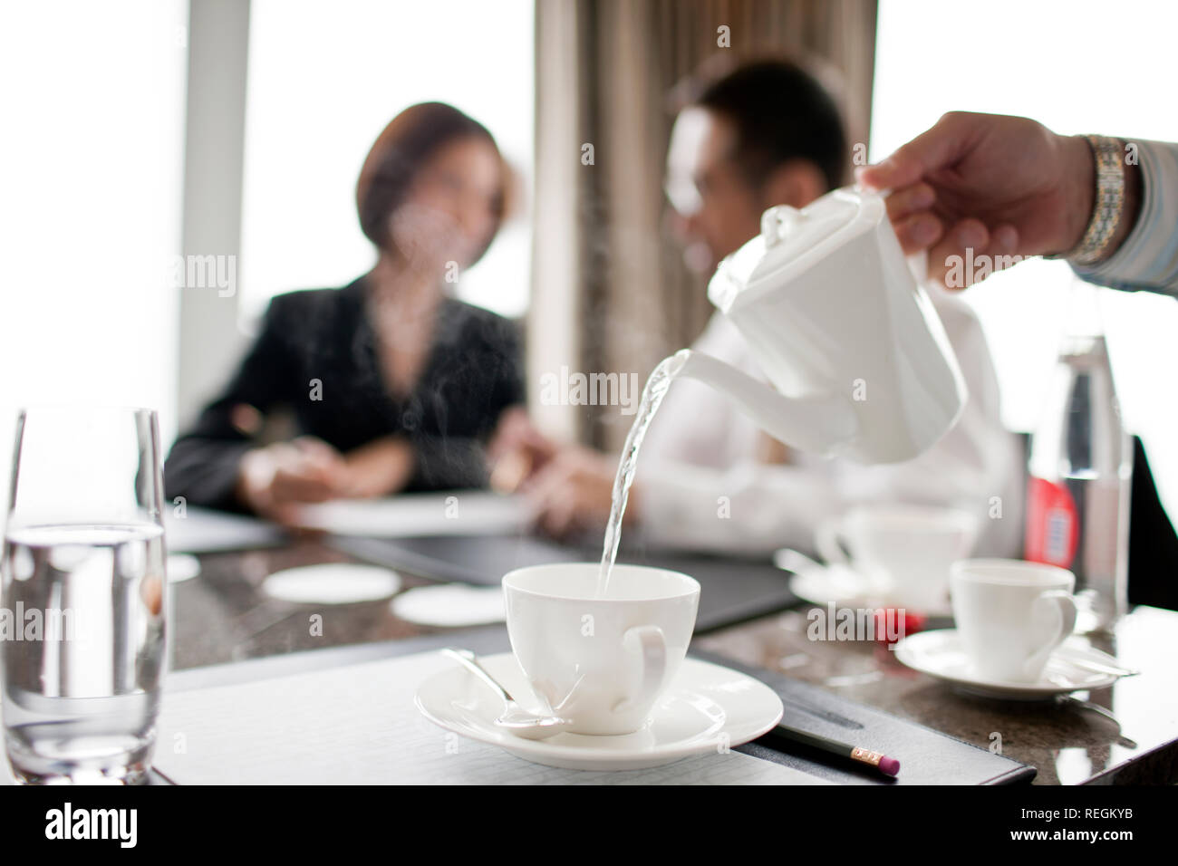 Businessman and woman talking at a conference table, with tea being poured in the foreground. Stock Photo