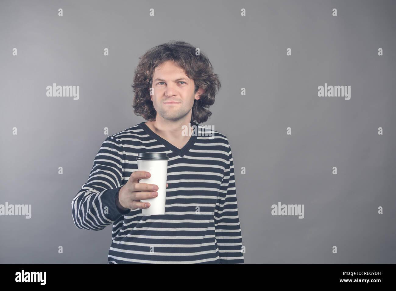 Portrait of happy handsome dark haired curly man in striped sweater with paper cup of morning coffee isolated on grey background. Good morning with te Stock Photo