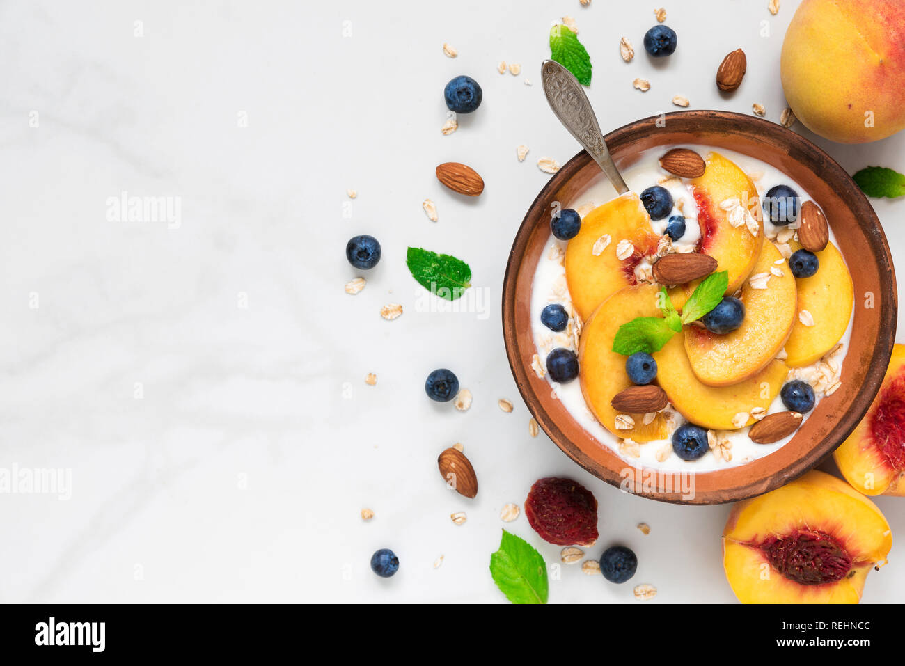 yogurt with peaches, oats, blueberries, honey, nuts and mint in a bowl with a spoon for healthy breakfast on white marble background. top view with co Stock Photo