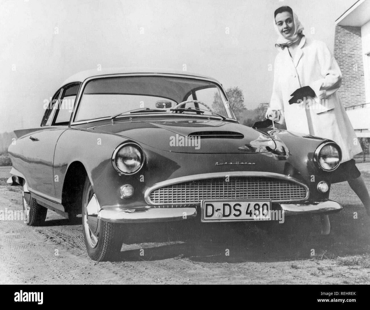 Driving in the 1950s. A young woman is standing beside a car Auto Union. She wears a typical 1950s short white coat with matching white scarf. 1950s Stock Photo