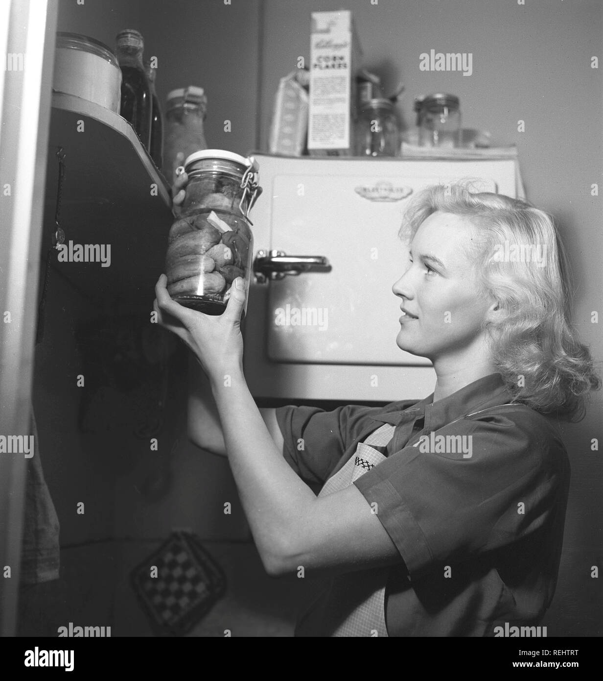 1940s practical tips. A young woman at home looks happy when holding a glass bottle with conserved cucumbers. She is in the kitchen dressed in an apron. The year is 1940 and the world war II is going on. Food and other necessities are rationed, therefore an own supply of food was neccesary.  Photo Kristoffersson 157-24 Stock Photo