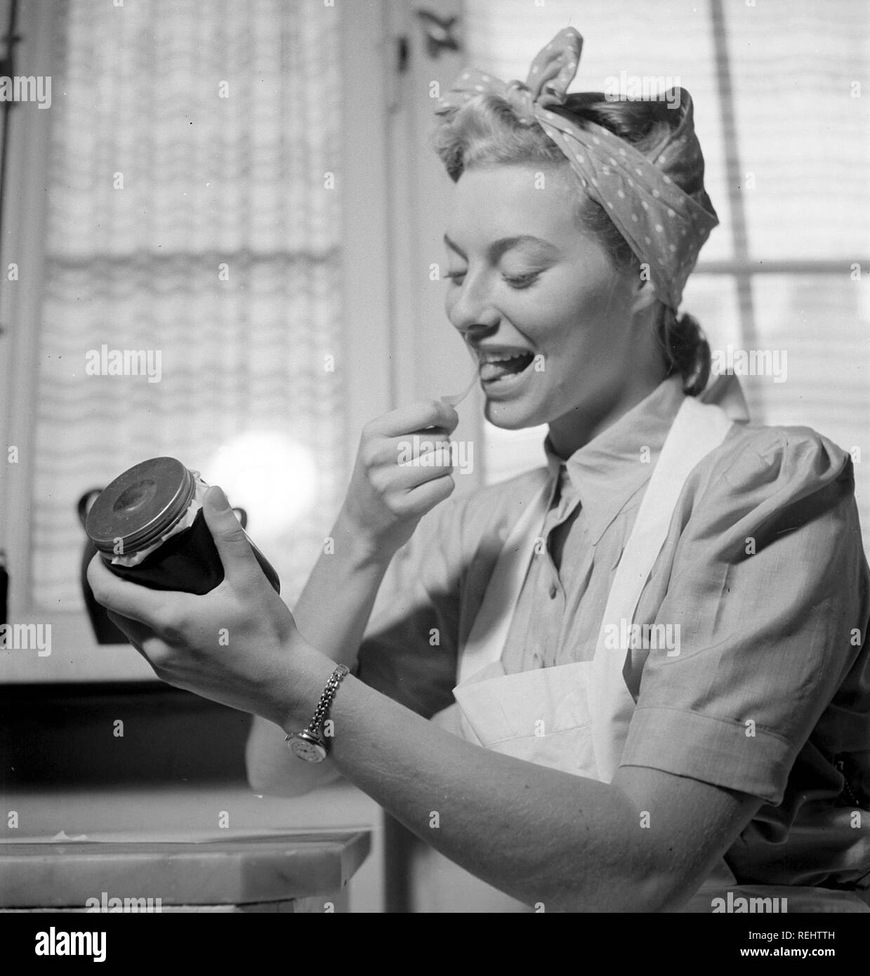 1940s practical tips. A young woman at home looks happy when licking the glue side of a label to put on the glass jar of the jam she has made herself. She is in the kitchen dressed in an apron and has her hair set up in a typical way. The year is 1940 and the world war II is going on. Food and other necessities are rationed, therefore an own supply of food was neccesary.  Photo Kristoffersson 158-1 Stock Photo