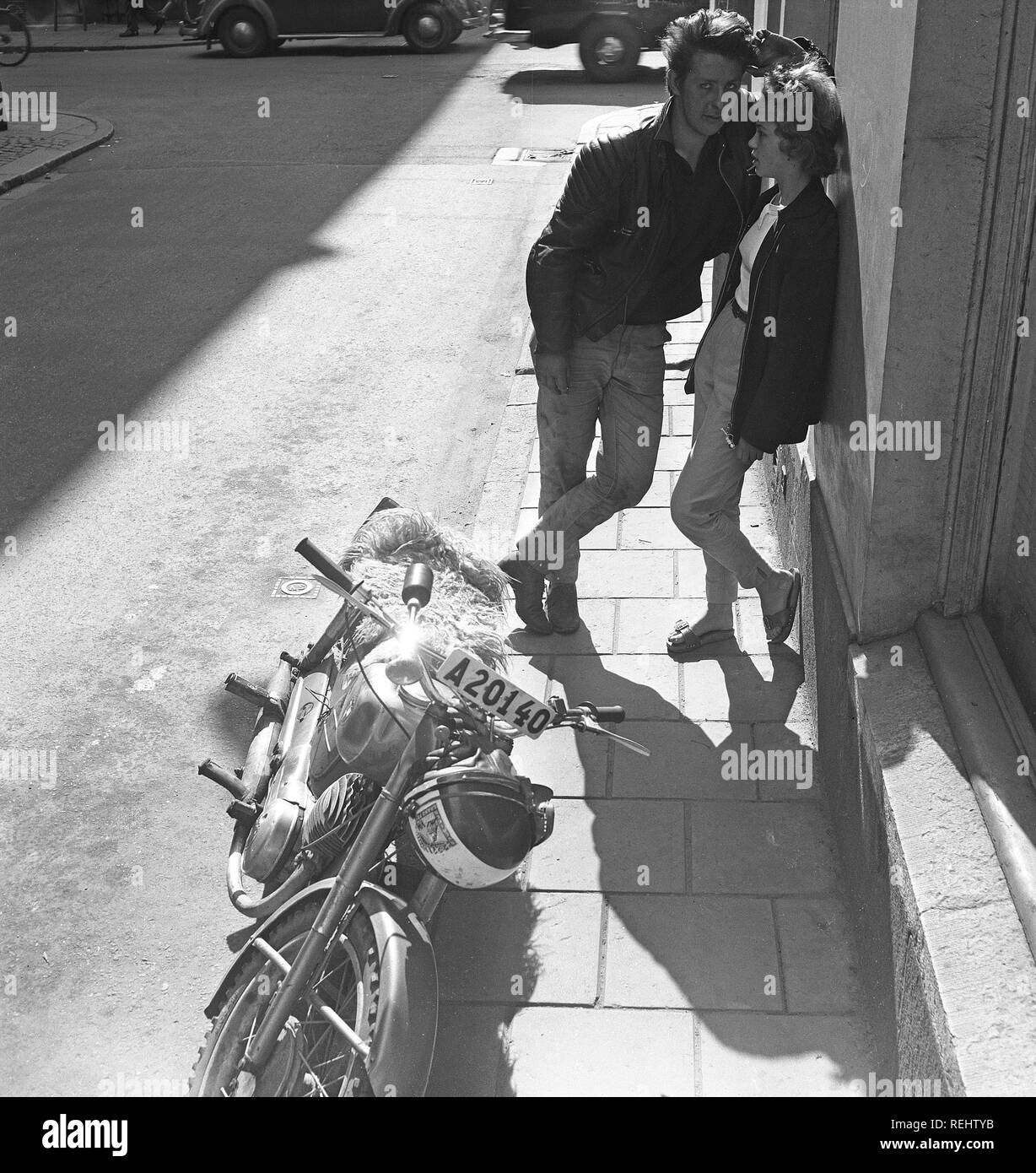 Couple in the 1950s. A young couple is standing leaning against a house wall in a Stockholm street. He has a Triumph motorcycle parked beside with a typical woolen padded seat. Photo Kristoffersson ref CA29-10. Sweden 1958 Stock Photo