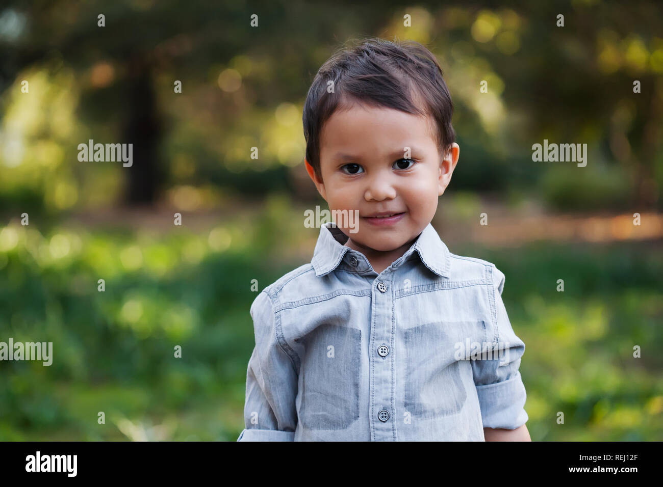 A cute mexican boy standing in a green wooded field, with gentle smile on his face and wearing a long sleeve shirt. Stock Photo