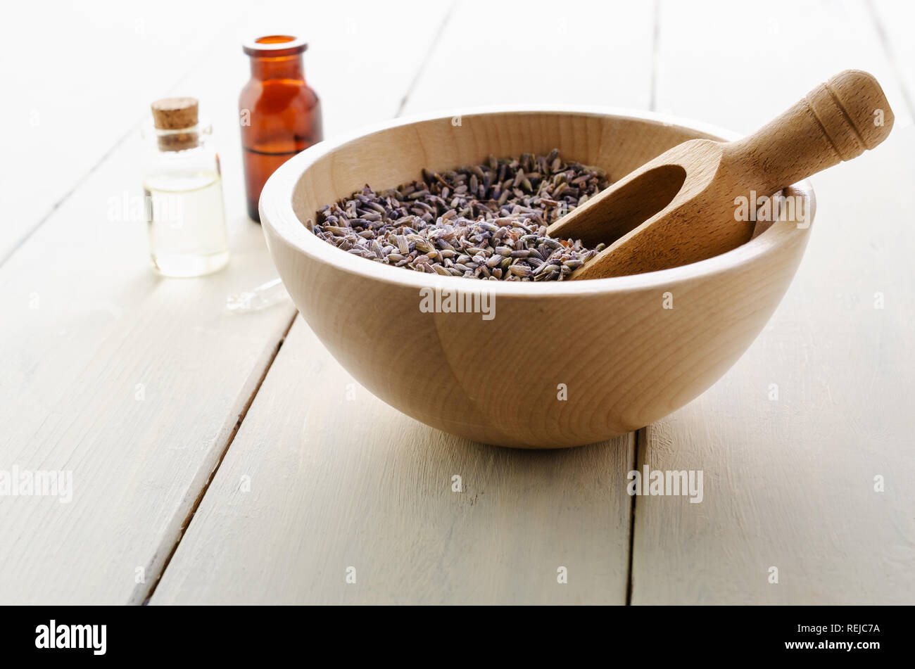 Herbal Lavender flower buds fill a wooden bowl with scoop in front of glass vials containing oils with pipette on white painted wood plank table. Stock Photo