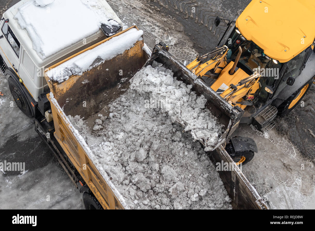 Tractor loader machine uploading dirty snow into dump truck. Cleaning city street, removing snow and ice after heavy snowfalls and blizzard Stock Photo
