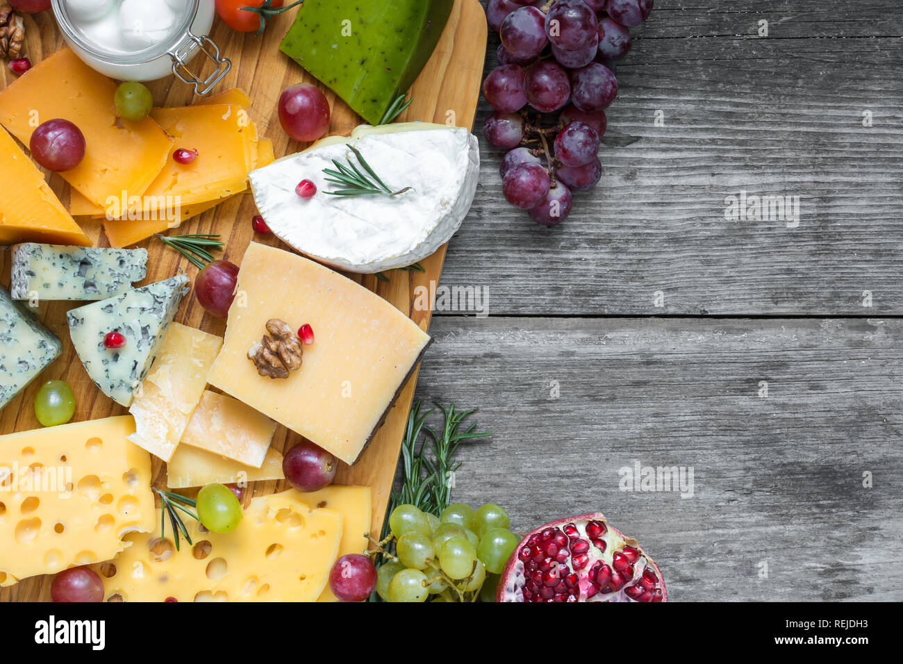 Assorted cheeses on wooden board plate served with walnuts, grapes, pomegranate and rosemary on rustic wood background, top view with copy space Stock Photo