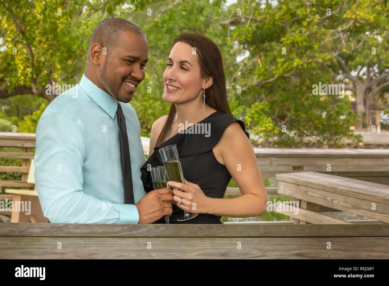 A modern interracial couple stands on an outdoor deck holding champagne glass laughing with joy. The couple faces each other with warm smiles. Stock Photo