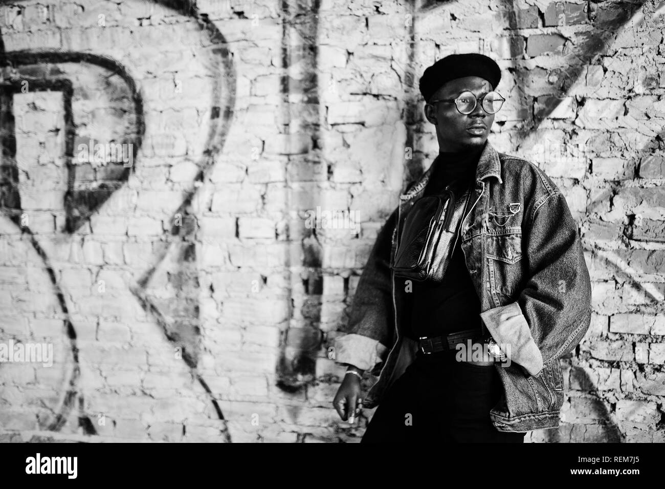 African american man in jeans jacket, beret and eyeglasses against graffiti brick wall with bruk sign. Stock Photo