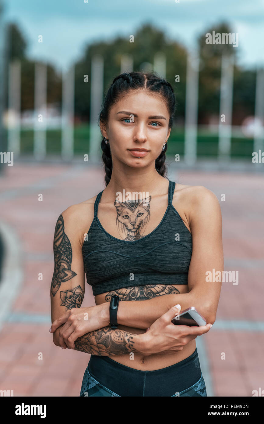 Beautiful girl is athlete with tanned skin and cat tattoos, in summer city.  Emotionally and confidently looks into frame. Concept of strength and  confidence. In sportswear with clock and smartphone Stock Photo -