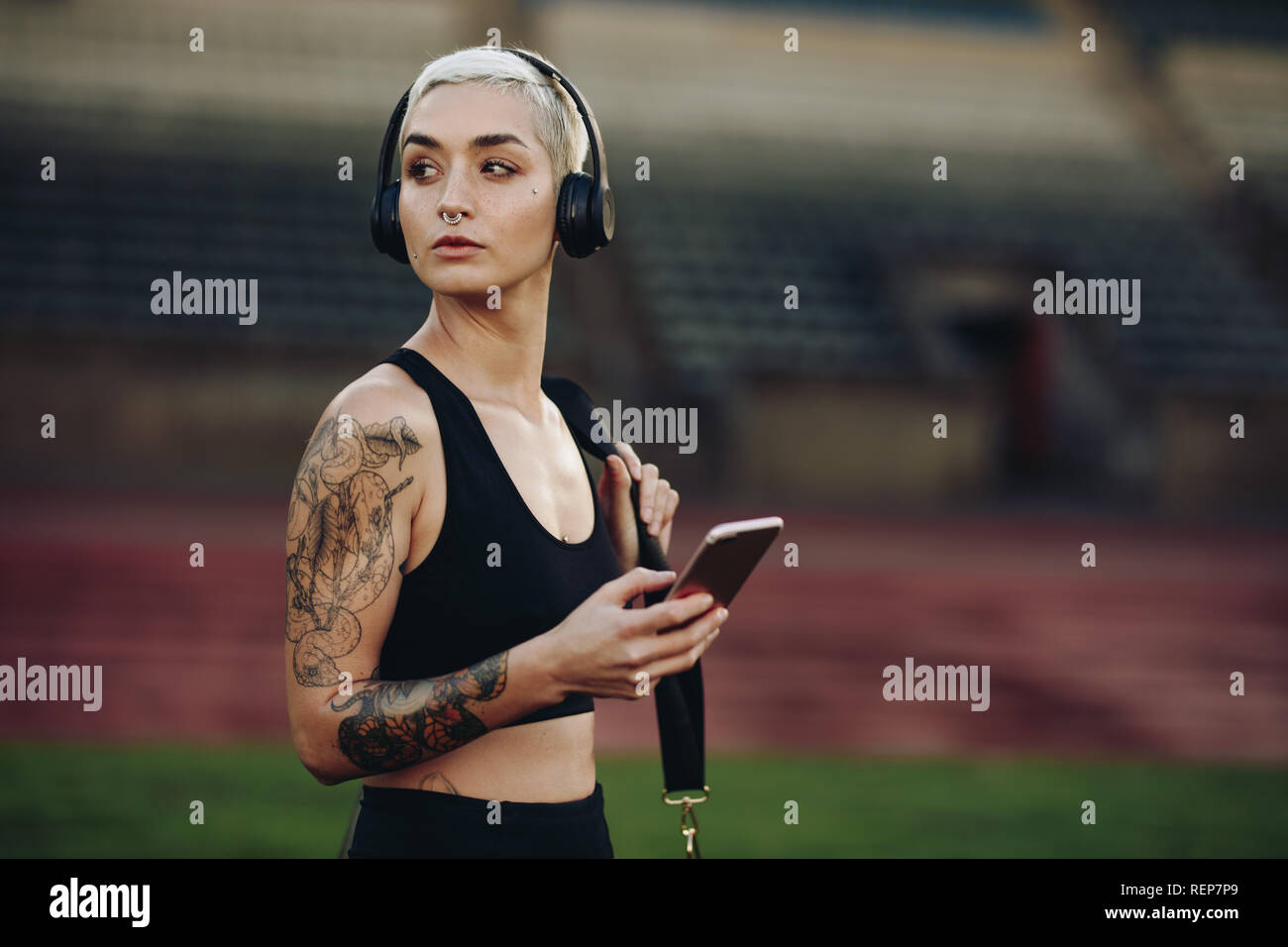 Side view of a female runner standing inside a track and field stadium carrying her bag. Female athlete listening to music on wireless headphones hold Stock Photo