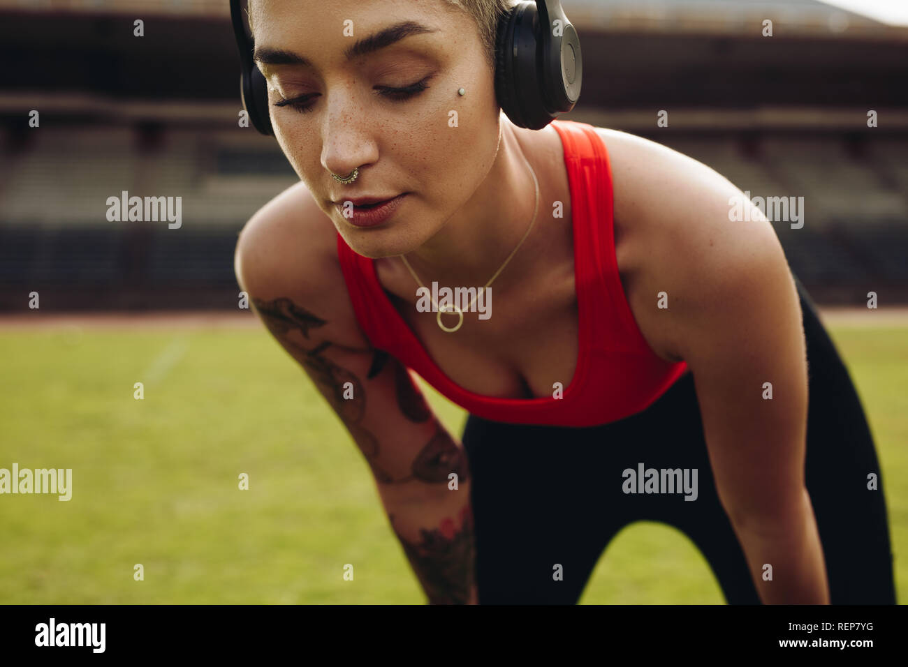 Close up of a woman athlete training in a track and field stadium with hands on knees. fitness woman wearing wireless headphones doing workout. Stock Photo