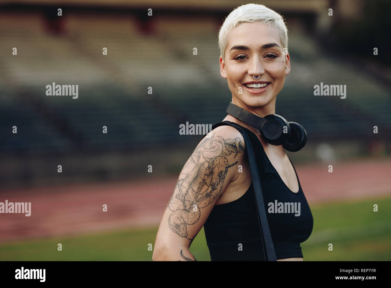 Cheerful fitness woman inside an athletic stadium with her bag and wireless headphones. Side view of a female athlete standing in a track and field st Stock Photo