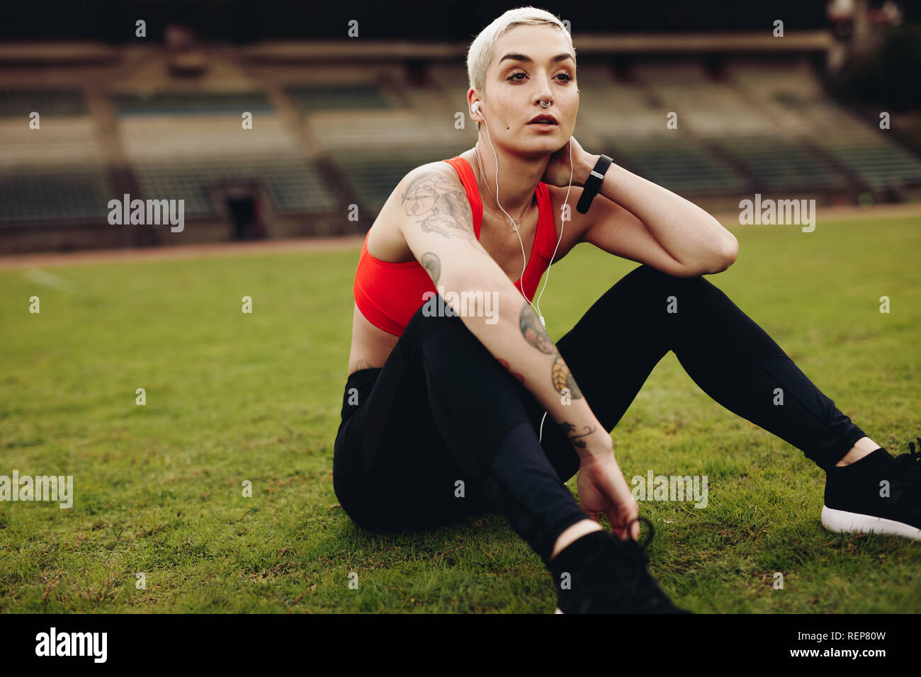 Female athlete sitting on grass listening to music on earphones. Woman runner relaxing after her workout in a track and field stadium. Stock Photo