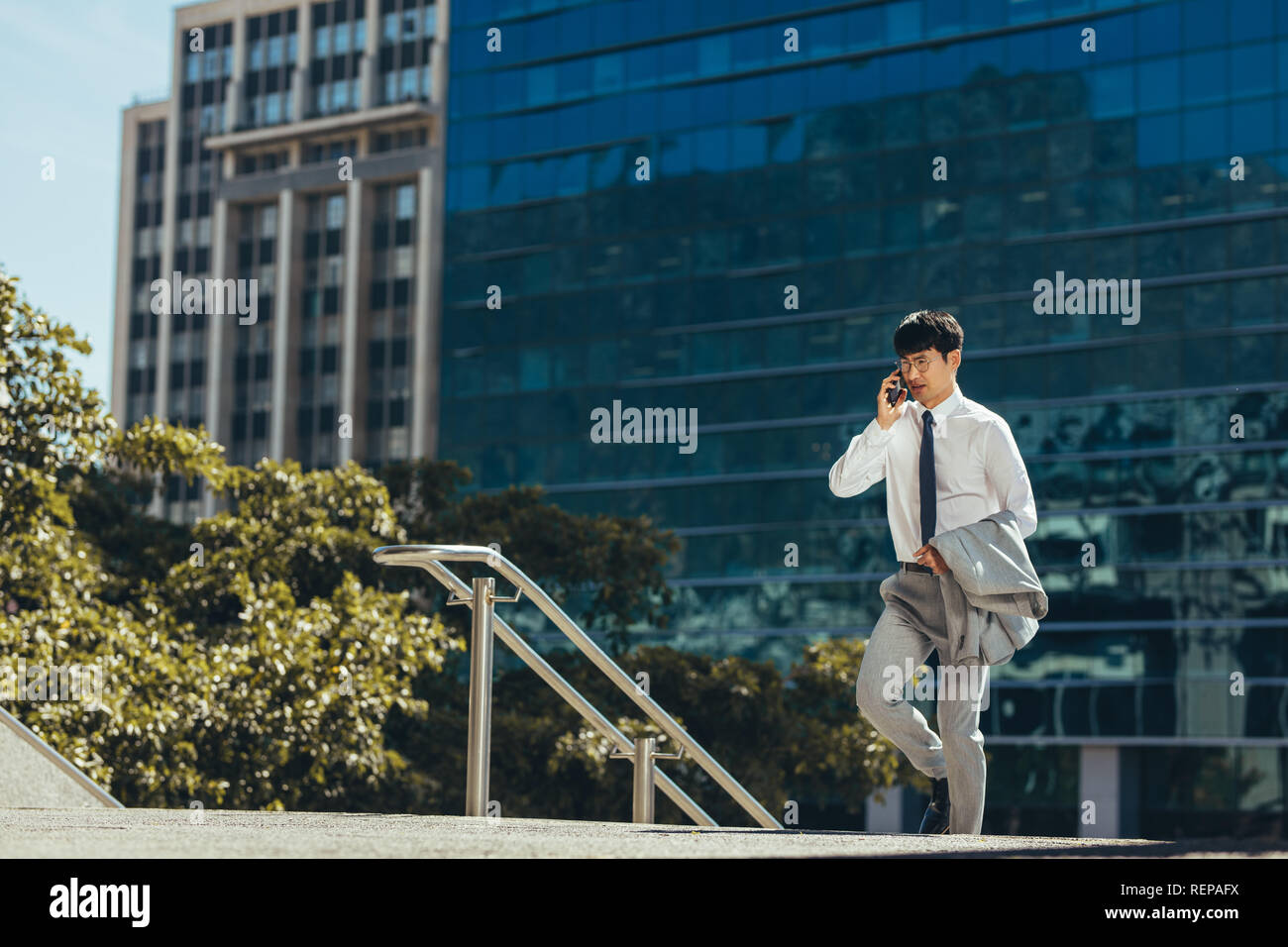 Asian businessman talking on his mobile phone outdoors while walking. Man in businesswear walking up the steps making a phone call against a office bu Stock Photo