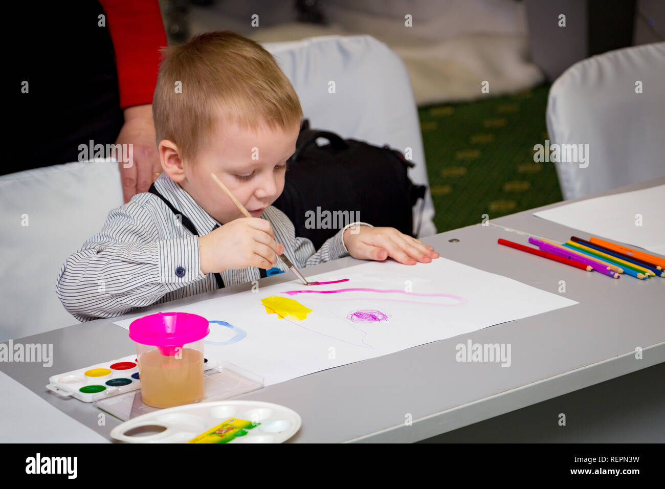 A little boy learns to paint with a brush on a piece of paper sitting at a table Stock Photo