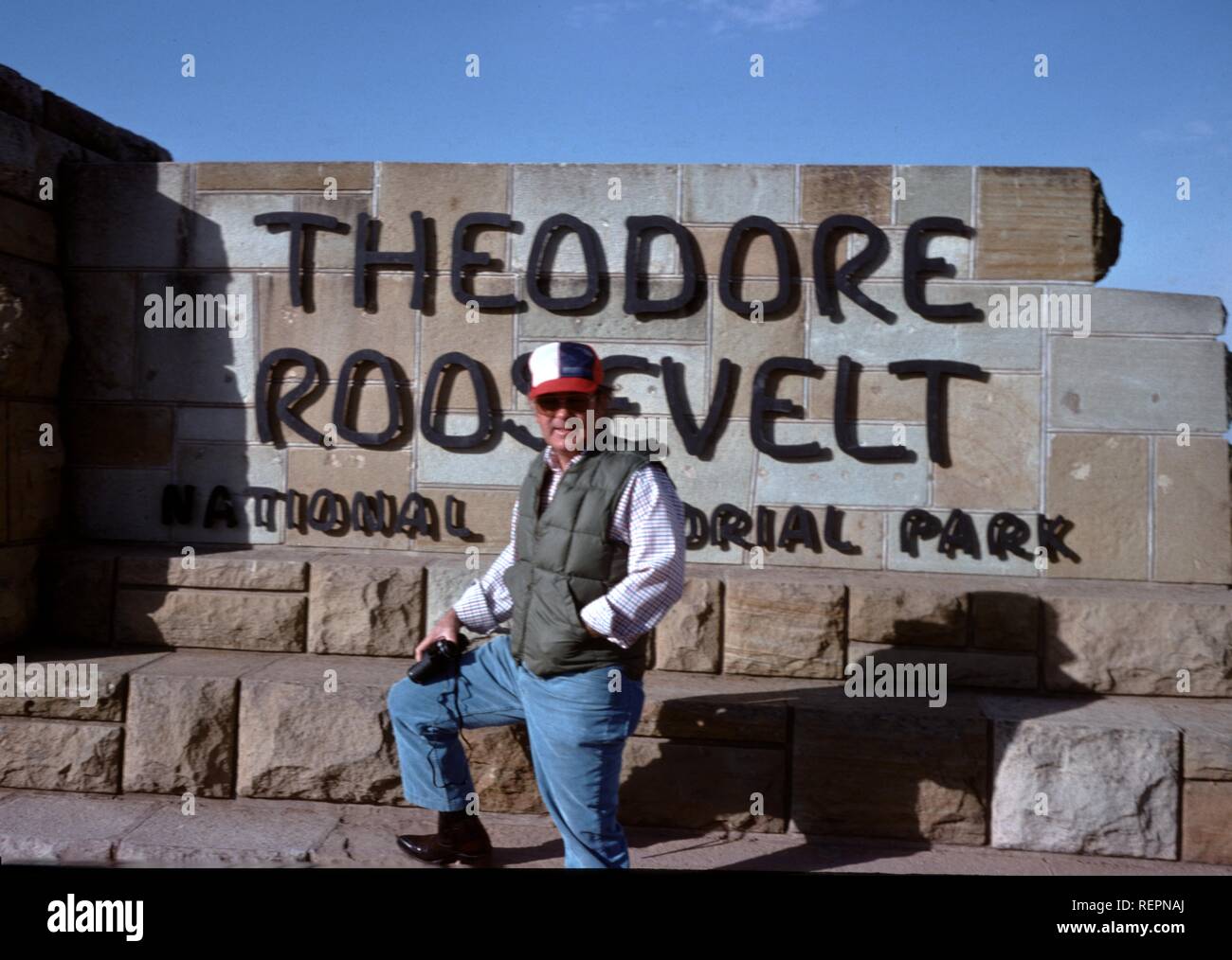 A man wearing a vest and hat and holding a camera stands with one leg up on the sign for Theodore Roosevelt National Memorial Park in Medora, North Dakota, 1976. () Stock Photo