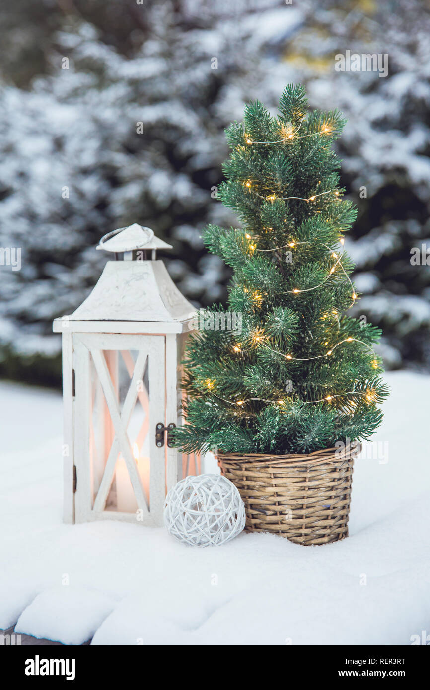 White wooden lantern with white wax candle lit and small Christmas fir tree with micro led lights in brown rattan flower pot in snow, snowy fir trees Stock Photo