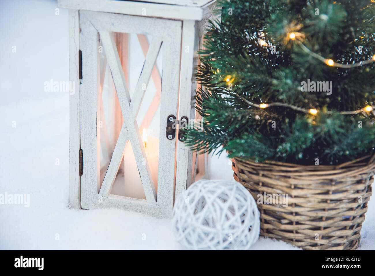 White wooden lantern with white wax candle lit and small Christmas fir tree with micro led lights in brown rattan flower pot in snow, snowy fir trees Stock Photo