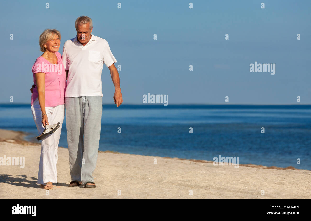 Happy senior man and woman couple walking laughing on vacation on a deserted tropical beach with bright clear blue sky and calm sea Stock Photo