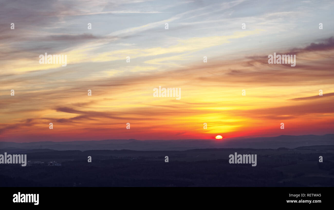 Colorful sunset in shades of yellow, red and orange with picturesque veil clouds in a hilly landscape, wide view Stock Photo