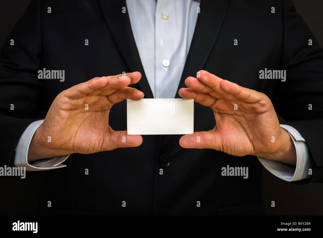 Man wearing a suit holding white business card on black wall background. Mock up, space for text Stock Photo