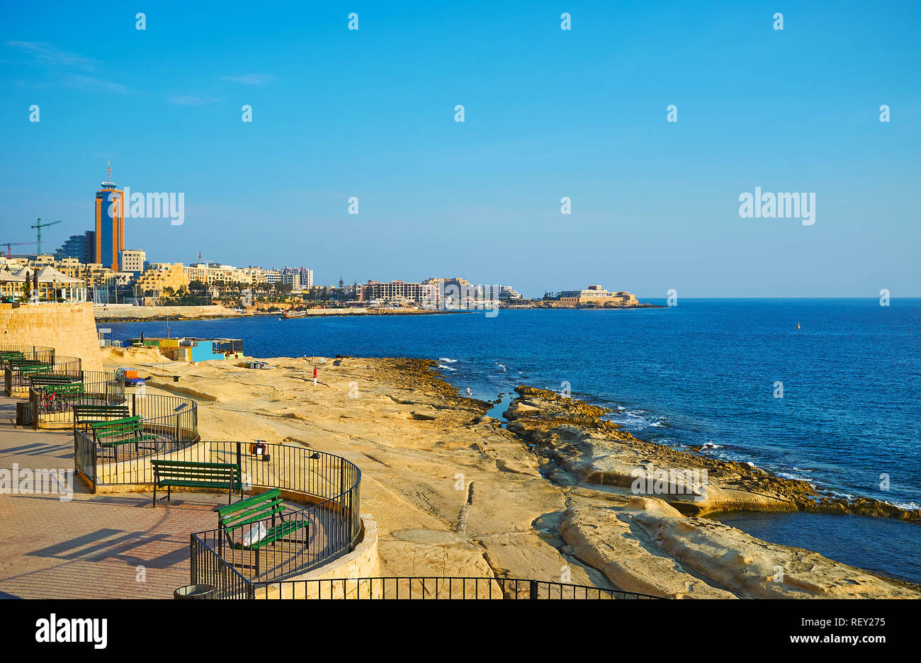 The nice seaside recreation area with benches on the rocky coast of Sliema with a view on the modern buildings of St Julians, Malta. Stock Photo
