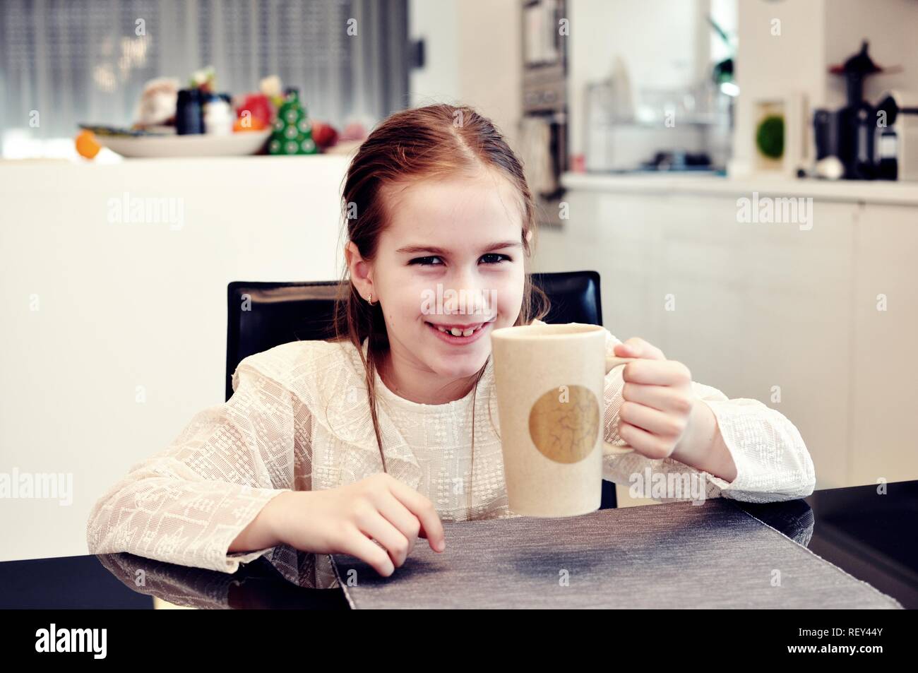 White Caucasian girl (child, kid) siting at the table, laughing and drinking a big cup of milk Stock Photo