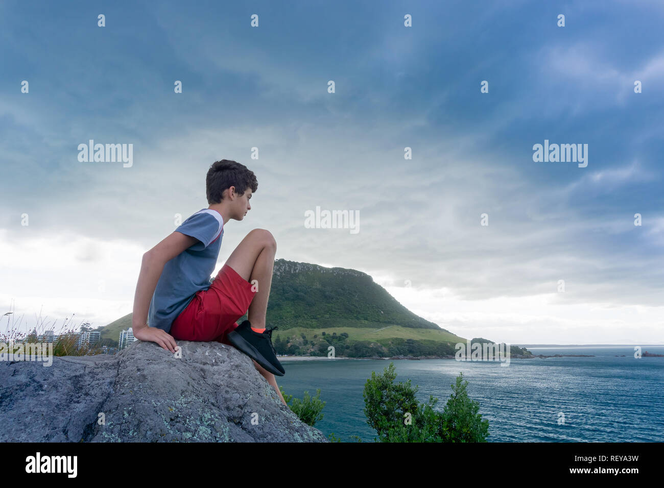 Boy sits on rock on Leisure Island looking out to sea with Mount Maunganui  under evening light and dark clouds in background Stock Photo