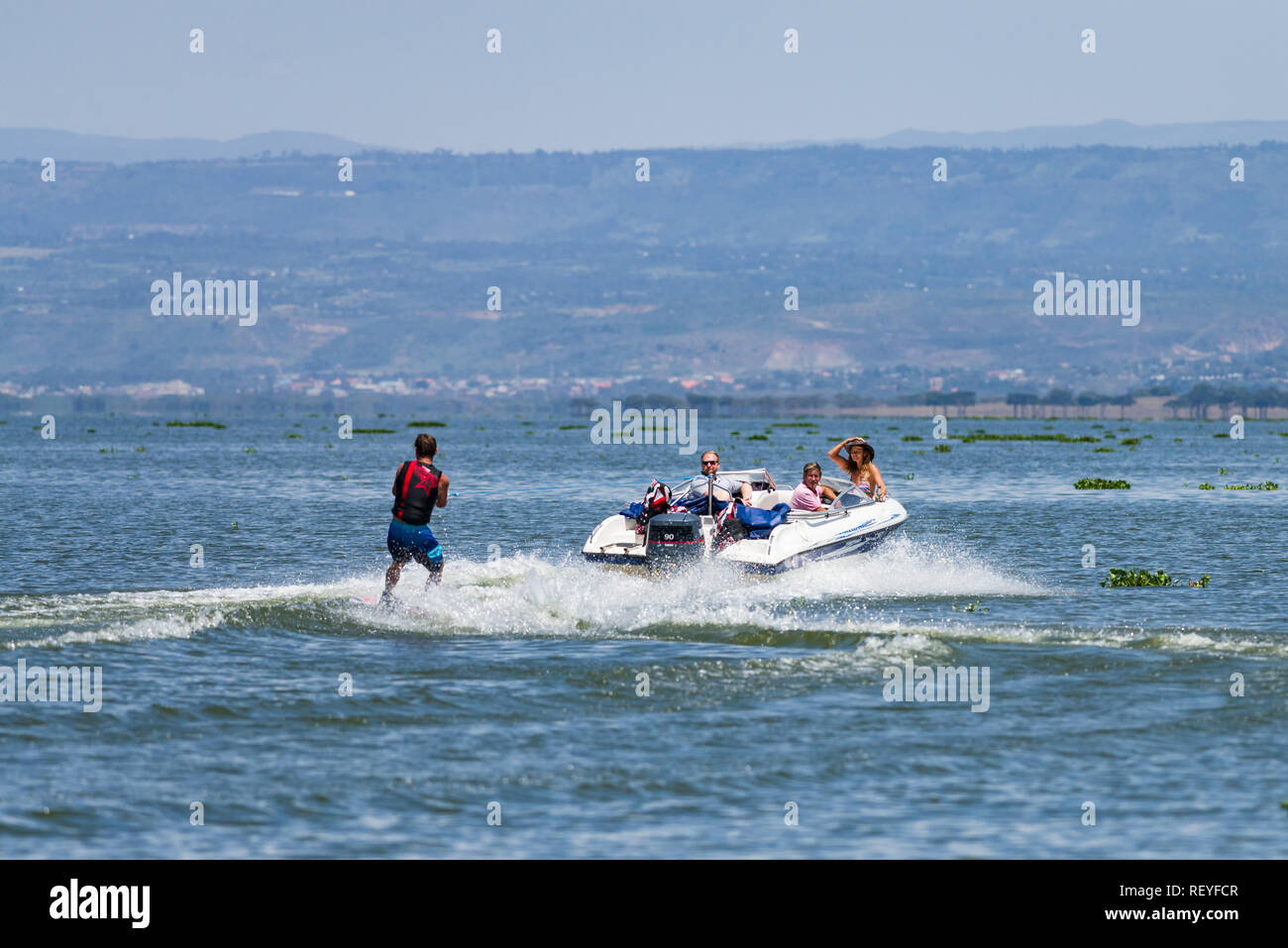 A Caucasian man wakeboarding behind a powerboat as passengers watch him, Lake Naivasha, Kenya Stock Photo