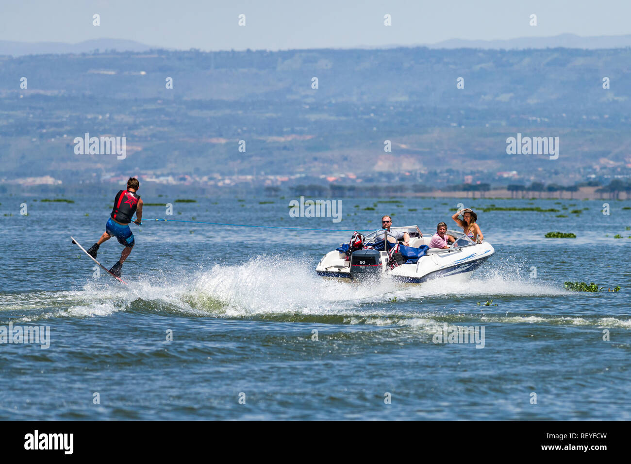 A Caucasian man performing a jump whilst wakeboarding behind a powerboat as passengers watch him, Lake Naivasha, Kenya Stock Photo