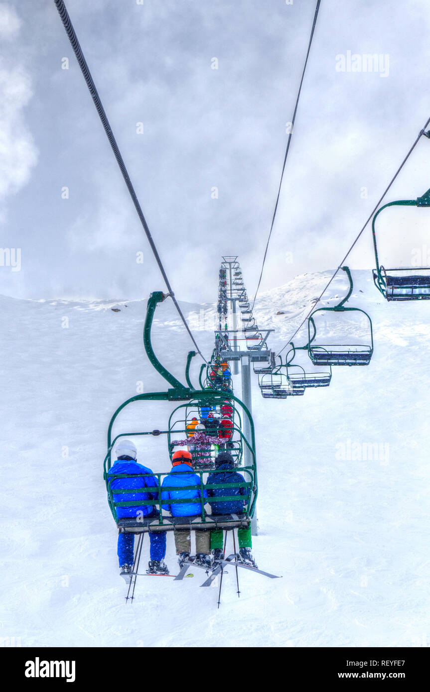 Unidentifiable skiers on chairlift going up a ski slope in the snowy mountain range of the Canadian Rockies. Stock Photo
